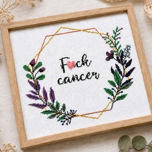 Pode incluir: Um bordado em ponto cruz quadrado numa moldura de madeira clara com uma borda geométrica dourada, ramos botânicos verdes e roxos, e o texto preto "Fuck cancer" com um pequeno coração rosa a substituir a letra u.