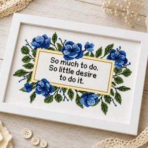 May include: White framed cross-stitch art with blue floral design. The artwork features the text "So much to do. So little desire to do it." The frame is rectangular and the artwork is surrounded by green leaves.