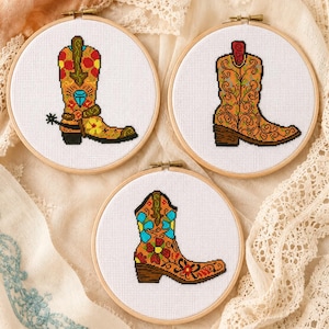 May include: Three circular wooden embroidery hoops containing white fabric with cross-stitch designs of cowboy boots. The top left boot features red, yellow, and blue floral patterns with a spur. The top right boot has orange scrollwork. The bottom boot displays blue and red floral motifs.