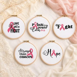 Könnte beinhalten: Fünf runde Stickrahmen aus Holz mit weißem Stoff und Kreuzstichmotiven. Die Designs zeigen rosa Schleifen zur Sensibilisierung und schwarzen Text: Live Love Fight, Pink is my power Color, Fight, Stronger than cancer und Hope.