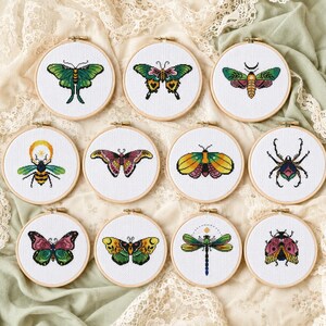 Puede incluir: Diez bastidores de bordado circulares de madera que contienen diseños de punto de cruz de varios insectos, incluyendo mariposas, polillas, una abeja, una araña, una libélula y un escarabajo, centrados en tela blanca con hilos de colores.