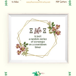 May include: Cross stitch pattern featuring a gold geometric frame with pink flowers and the text "X Life X is just a random series of screwups on a countdown timer". The pattern is for Aida 14 count fabric and measures 11.6 by 10.2 inches (30.1 by 25.8 cm).