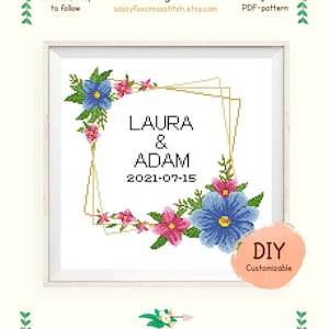 May include: Cross stitch pattern featuring a floral wreath with blue and pink flowers, a gold geometric frame, and the text "LAURA & ADAM 2021-07-15". The pattern is for Aida 14 count fabric and uses 23 colors from the DMC palette.