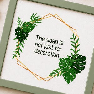 Könnte beinhalten: Ein quadratisches Kreuzstichbild in einem salbeigrünen Rahmen mit einer goldenen, geometrischen Sechseck-Kontur, umgeben von grünen Monstera- und Farnblättern. Der mittlere Text lautet, The soap is not just for decoration, in schwarzer Schrift.