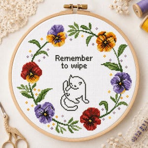 May include: Cross-stitch embroidery in a wooden hoop featuring a floral wreath of pansies in shades of purple, yellow, and red. The text "Remember to wipe" is above a line drawing of a cat. A charming, handmade piece.