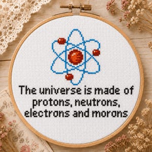 May include: A cross-stitch embroidery hoop featuring a blue and red atom design. The text reads, "The universe is made of protons, neutrons, electrons and morons." The embroidery is set in a wooden hoop.