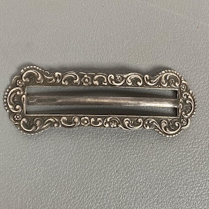 Pode incluir: Uma fivela de metal prateado de formato retangular com uma borda em relevo adornada com padrões florais e volutas intrincados. A fivela possui uma barra horizontal central e duas aberturas, com uma marca visível na superfície interna.