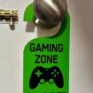 May include: Green door hanger with the text "Gaming Zone" and a black video game controller graphic. The text "Enter with Caution" is at the bottom of the hanger.