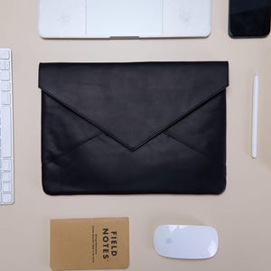 May include: A black leather envelope-style laptop sleeve with a pointed flap closure, positioned centrally among a white keyboard, a white computer mouse, a tan Field Notes notebook, a white stylus, a smartphone, and a laptop computer.