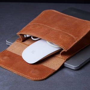 Könnte beinhalten: Eine braune Leder-Maus-Hülle mit einer weißen Maus im Inneren. Die Hülle ist geöffnet und zeigt die Maus und ein Ladekabel. Die Hülle liegt auf einem Laptop.