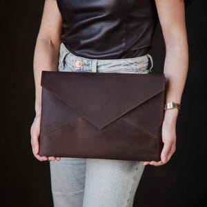 May include: A dark brown leather envelope-style clutch bag with a pointed flap closure and visible stitching along the edges. The rectangular bag features a smooth, matte finish and a clean, minimalist design suitable for carrying documents or items.