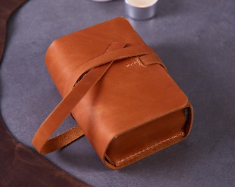 May include: A rectangular tan leather journal featuring a wrap-around leather strap closure. The cover is constructed from a single piece of smooth, matte-finish leather with visible stitching details along the edges and a secure tie mechanism.