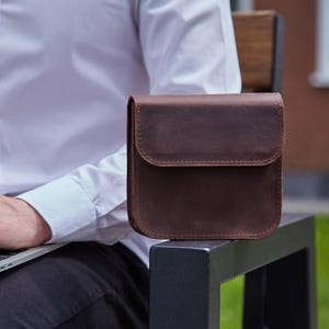 May include: A square-shaped dark brown leather bag with a fold-over flap and visible stitching along the edges. The bag features a smooth, matte finish and a structured design, presented as a compact accessory for carrying small personal items.