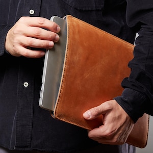 Puede incluir: Una funda de cuero color canela para portátil con costuras visibles en los bordes, que contiene un portátil plateado. La funda está fabricada con un material liso de acabado mate y presenta un diseño sencillo con la parte superior abierta.