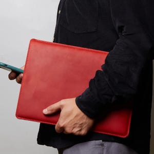 Puede incluir: Una funda rectangular de cuero rojo para computadora portátil con costuras visibles a lo largo de los bordes. La funda tiene un acabado liso y mate con un perfil delgado, diseñada para sostener una computadora portátil de forma segura.