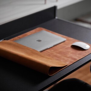 Può includere: Un tappetino da scrivania in pelle marrone con un bordo arrotolato, un laptop argentato e un mouse bianco su una scrivania nera. Il tappetino offre una superficie liscia per il laptop e il mouse.
