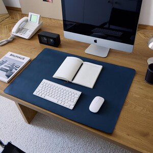 Puede incluir: Una alfombrilla de escritorio rectangular de color azul marino sobre una mesa de madera. La alfombrilla sirve de base para un teclado inalámbrico blanco, un ratón de ordenador blanco y un cuaderno abierto con páginas rayadas y un monitor.