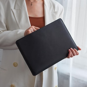 May include: A black rectangular laptop sleeve held by a person wearing a white blazer and a brown top. The sleeve appears to be made of leather or a similar material, with a smooth, matte finish. The background is a soft, neutral color.