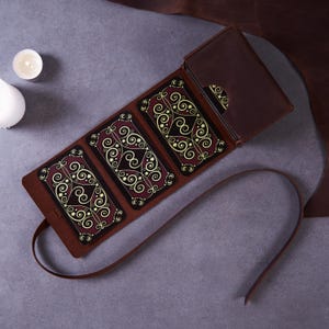May include: A brown leather tarot card holder with a flap closure, holding several tarot cards. The cards have a burgundy background with gold and cream-colored swirling designs. A leather strap extends from the holder.
