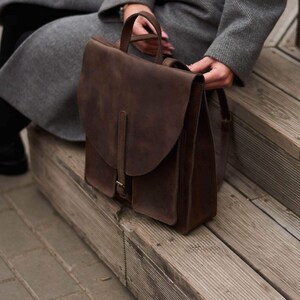 May include: A dark brown leather backpack featuring a large fold-over flap with a vertical strap and buckle closure. The bag has a top handle, two side pockets, and a structured rectangular body with a matte, smooth finish and visible stitching.