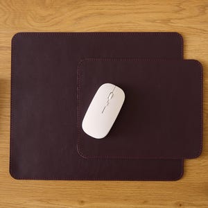Puede incluir: Una alfombrilla de escritorio rectangular de color morado oscuro con esquinas redondeadas y bordes cosidos, que presenta una alfombrilla más pequeña a juego superpuesta. Un ratón de ordenador inalámbrico blanco se sitúa sobre la alfombrilla pequeña, junto a un teclado blanco.