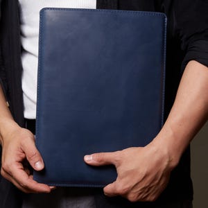 May include: A rectangular navy blue leather document holder or laptop sleeve with a smooth, matte finish and stitched edges. The item is held vertically, showcasing a clean, minimalist design with a solid color and a structured, flat rectangular shape.