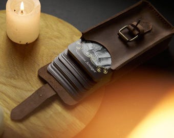 May include: A brown leather deck holder with a metal buckle closure containing a stack of rectangular cards. The top card features a black and white illustration with gold decorative flourishes and the printed text Nummus Armiger.