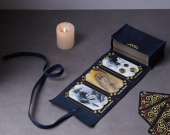 May include: A dark blue fabric tarot card case with a long tie closure, containing a deck of cards. Three cards are displayed face-up in a row, featuring illustrations and the text "Tres Numen", "Druida Tubulis", and "Benevoti Pugionibus".