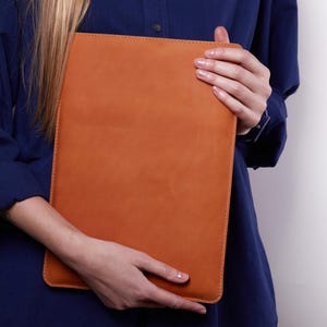 May include: A rectangular, tan-colored leather sleeve with visible stitching along the edges. The sleeve has a smooth, matte texture and a simple, flat design, providing a protective cover for a tablet or laptop device.