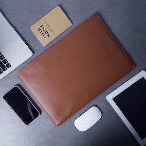 Pode incluir: Uma capa retangular de couro castanho para portátil posicionada ao centro, rodeada por um portátil prateado, um caderno Field Notes bege, um smartphone preto, um rato branco e um tablet branco sobre uma superfície cinzenta.