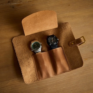 Op de afbeelding: Een horlogerol van bruin leer met twee binnenvakken, elk met een polshorloge. Het ene horloge heeft een zilverkleurige kast met een groene wijzerplaat, terwijl het andere een zwarte kast en band heeft. Voorzien van een klepsluiting met metalen gesp.