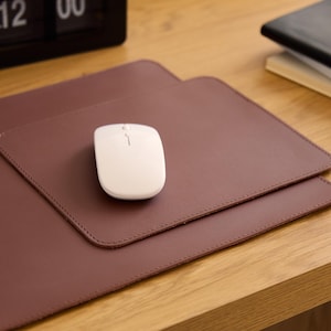 Può includere: Un tappetino da scrivania rettangolare color marrone scuro con superficie liscia, angoli arrotondati e cuciture visibili lungo i bordi. Un mouse per computer wireless bianco con rotellina centrale è posizionato al centro del tappetino.