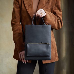 May include: A rectangular black leather tote bag with a vertical orientation, featuring a single exterior zippered pocket on the front and two integrated top handles. The bag has a smooth, matte finish and a clean, structured design.