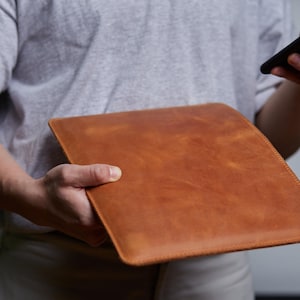 May include: A rectangular, tan-coloured leather sleeve with a smooth, matte finish and visible stitching along the edges. The sleeve is flat and features a simple, unadorned design with a consistent, warm brown tone throughout the surface.