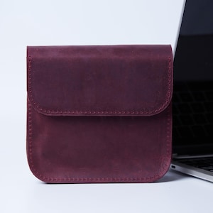 May include: A square-shaped, deep burgundy leather pouch featuring a fold-over flap with visible stitching along the edges. The pouch has a matte finish and a structured design, positioned next to the corner of a silver laptop on a white surface.
