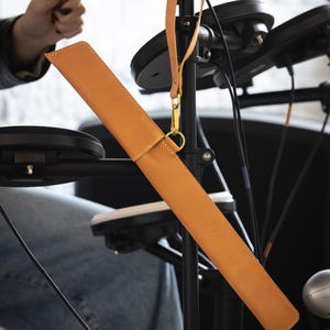 Op de afbeelding: Een bruinleren drumstokkenetui met een bijpassende riem en een goudkleurige sluiting. De etui is rechthoekig en heeft een gestikte rand. De achtergrond omvat een zwart elektronisch drumstel en een persoon die een drumstok vasthoudt.