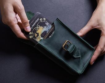 May include: A dark green leather pouch featuring a rectangular shape with visible stitching along the edges and a brass-toned metal buckle closure. A black card with gold decorative text reading Alchymista is partially inserted into the pouch.