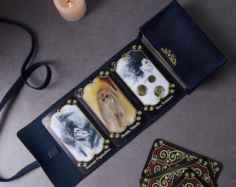 May include: A dark blue leather tarot card holder containing three displayed cards labeled Sexcenti Pugionibus, Druida Tubulis, and Tres Nummos, with a stack of cards inside the pocket and a matching leather tie strap attached to the side.