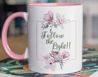 May include: White ceramic mug with a pink rim and handle. The mug features a floral design with pink flowers and a bird. The text "Follow the Light!" is printed on the mug.