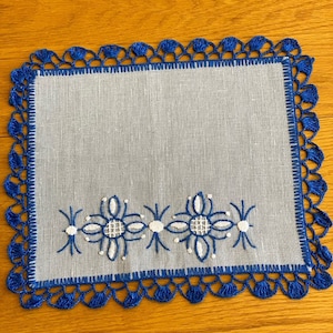 Könnte beinhalten: Eine weiße Leinenserviette mit blauer Häkelkante und gestickten weißen Blumen. Die Serviette liegt auf einer Holzoberfläche.