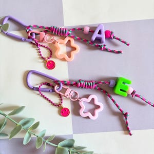 May include: Two keychains with purple carabiner clips, colourful cords, and letter charms. One has a pink "A" and the other a green "E". Each has a star-shaped charm and a smiley face charm. The keychains are on a checkered background.