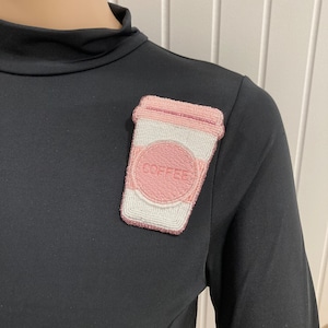 May include: A pink and white beaded coffee cup pin with the word "COFFEE" embroidered on the front. The pin is attached to a black long-sleeve shirt.