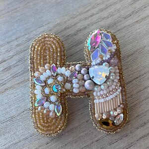 Puede incluir: Broche artesanal con forma de letra "H", adornada con cuentas. La base es dorada, con cristales iridiscentes, perlas y cuentas en tonos blanco, rosa y dorado. Los bordes están delineados con cuentas doradas, creando un accesorio decorativo.