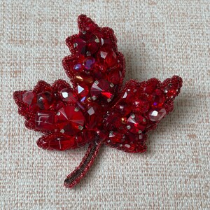 Puede incluir: Broche de hoja roja hecha a mano con cuentas. El broche tiene forma de hoja de arce, con intrincados abalorios y brillantes cristales rojos. El tallo es de color marrón oscuro.