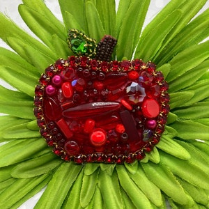 Puede incluir: Un broche de manzana roja hecho de cuentas de vidrio y diamantes de imitación. La manzana está rodeada de tela verde.
