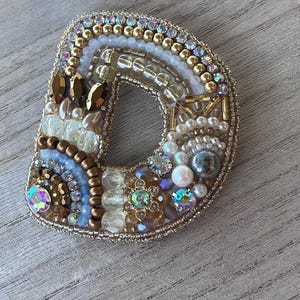 May include: A decorative brooch in the form of the letter "D", intricately adorned with beads, pearls, and crystals. The brooch has a gold-coloured outline and a spectrum of colours, including white, gold, blue, and iridescent highlights.