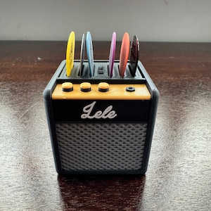 May include: A black and yellow guitar pick holder shaped like a small amplifier. The holder has six slots for guitar picks and the word "Lele" is printed on the front.