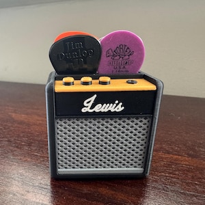 May include: A small black and yellow guitar amplifier with the word "Lewis" printed on the front. Three guitar picks are on top of the amplifier, two are black with white text and one is purple with white text.
