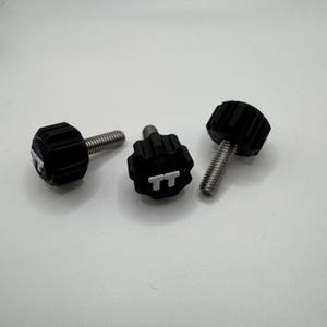 May include: Three black thumb screws with silver threaded shafts are arranged on a white surface. The screw heads have a ridged design for grip. One screw head has the letters "TT" in white.