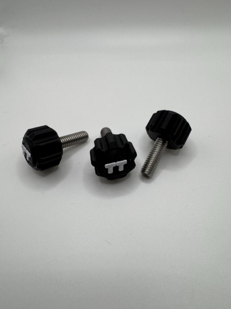 May include: Three black thumb screws with silver threaded shafts are arranged on a white surface. The screw heads have a ridged design for grip. One screw head has the letters "TT" in white.